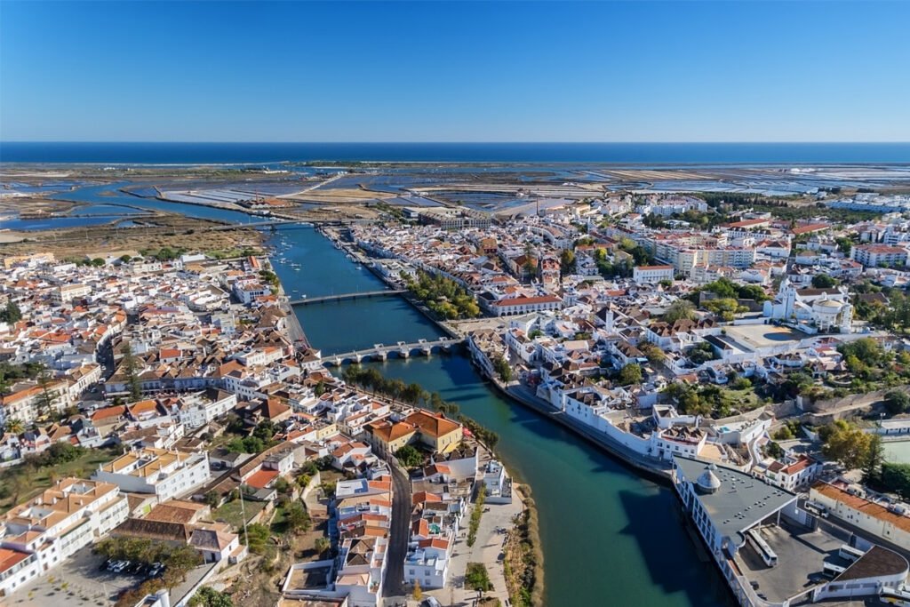 Aerial panoramic view of Tavira city in Algarve, Portugal, showing the Gilão River, the Roman Bridge, and the surrounding salt pans under a clear blue sky