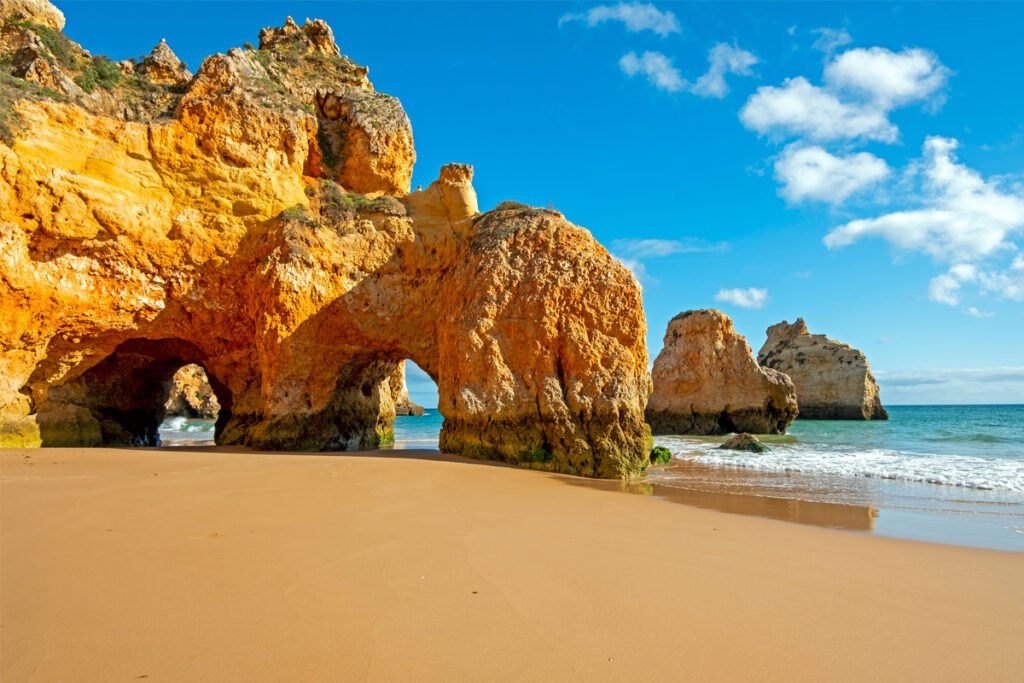 Panoramic photo of Praia da Marinha in Lagos, Algarve, featuring massive golden limestone sea arches and a secluded sandy beach under a clear blue sky with turqouise water