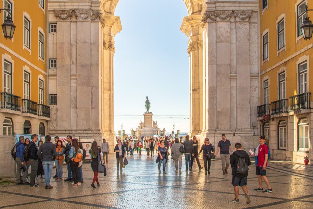 A perspective view through the monumental stone arch of Rua Augusta towards the Praça do Comércio in Lisbon, showing the statue of King José I in the distance, yellow buildings, and people walking on a traditional Portuguese pavement