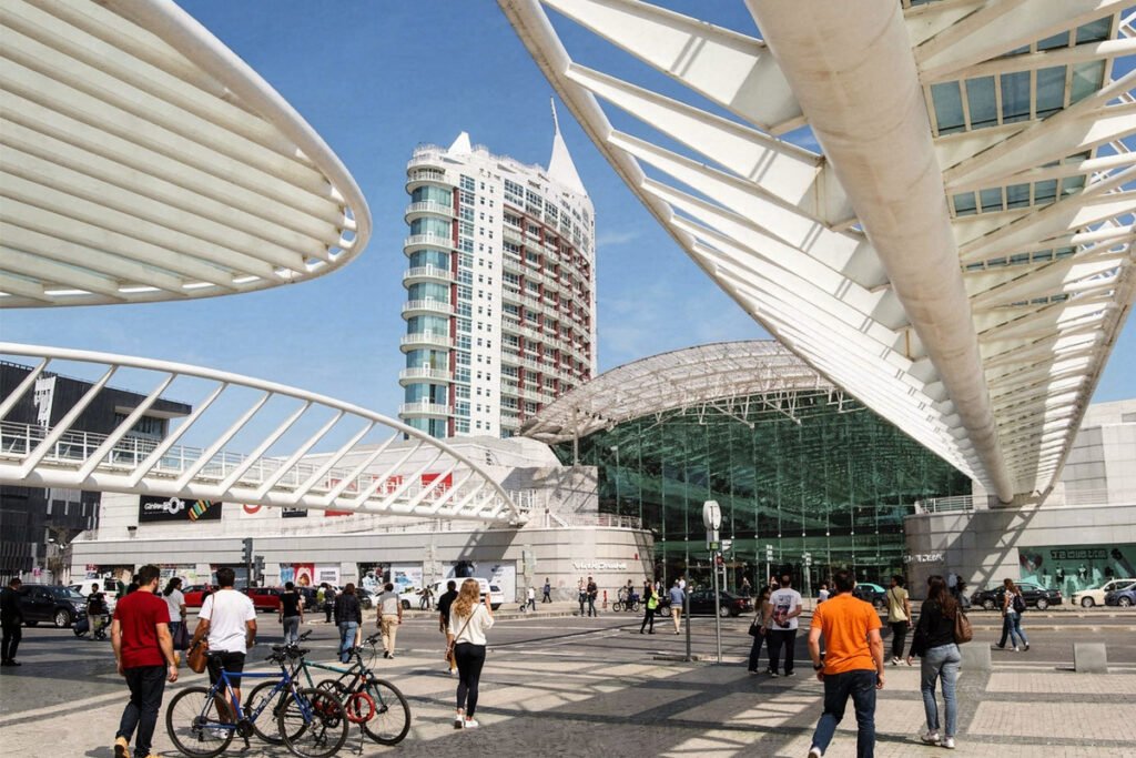 A wide shot of the modern Parque das Nações district in Lisbon, featuring the futuristic white steel canopies of the Gare do Oriente station, a tall contemporary residential building, and people walking on a sunny day