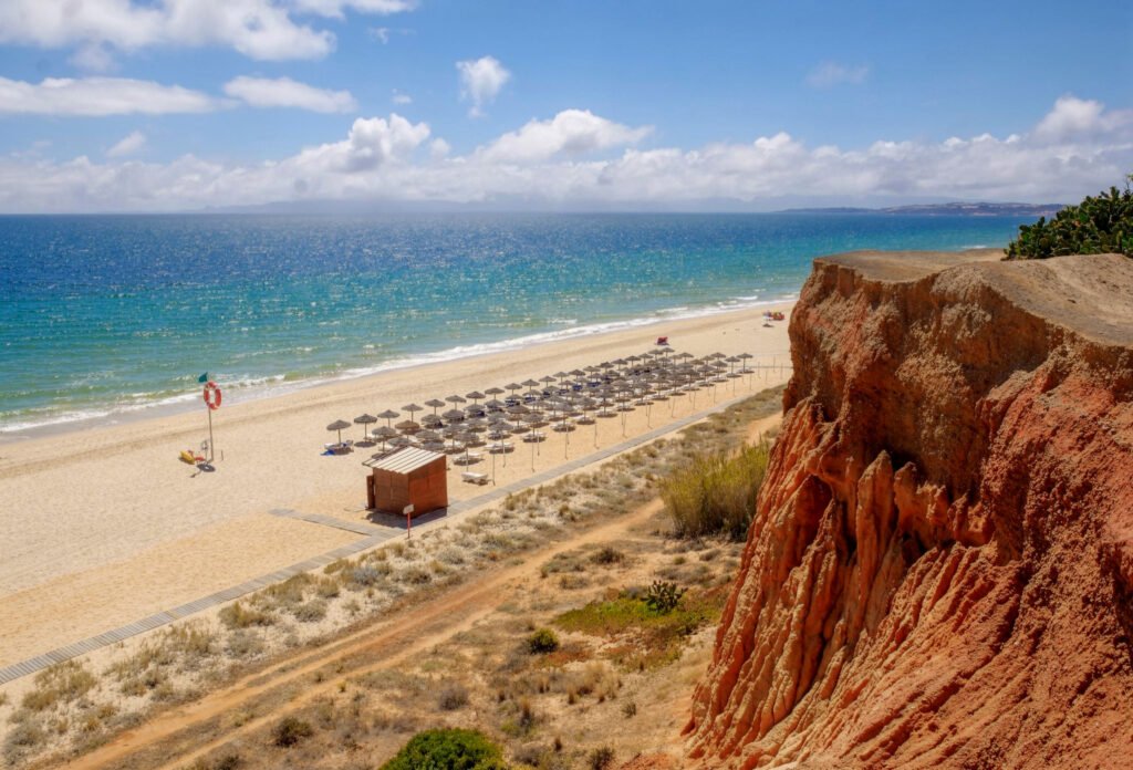 A panoramic high-angle view of Praia da Falésia in Algarve, Portugal, featuring its signature red and orange sandstone cliffs, a long golden sand beach with straw umbrellas, and the turquoise Atlantic Ocean under a blue sky with white clouds