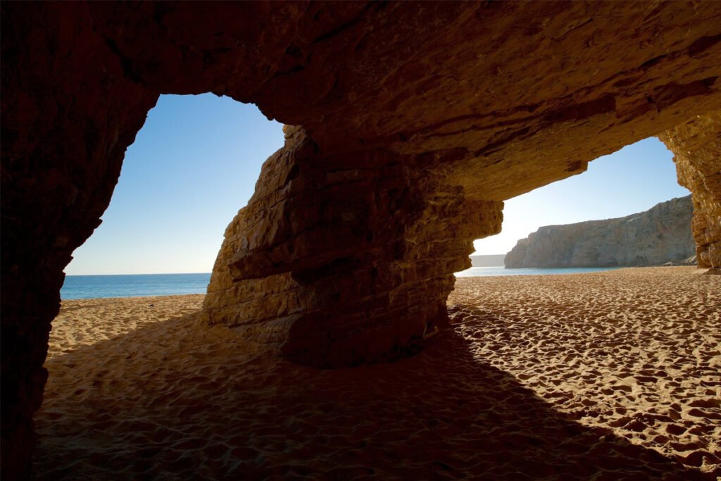View from inside a large sea cave at Praia do Beliche in Sagres, showing the sandy beach, high cliffs, and the deep blue Atlantic Ocean