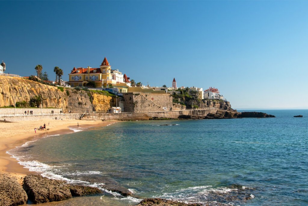 A scenic view of Tamariz Beach in Estoril, Portugal, showing the turquoise ocean, golden sand, and the famous Chalet Barros castle on the cliffs, located along the coast toward Cascais