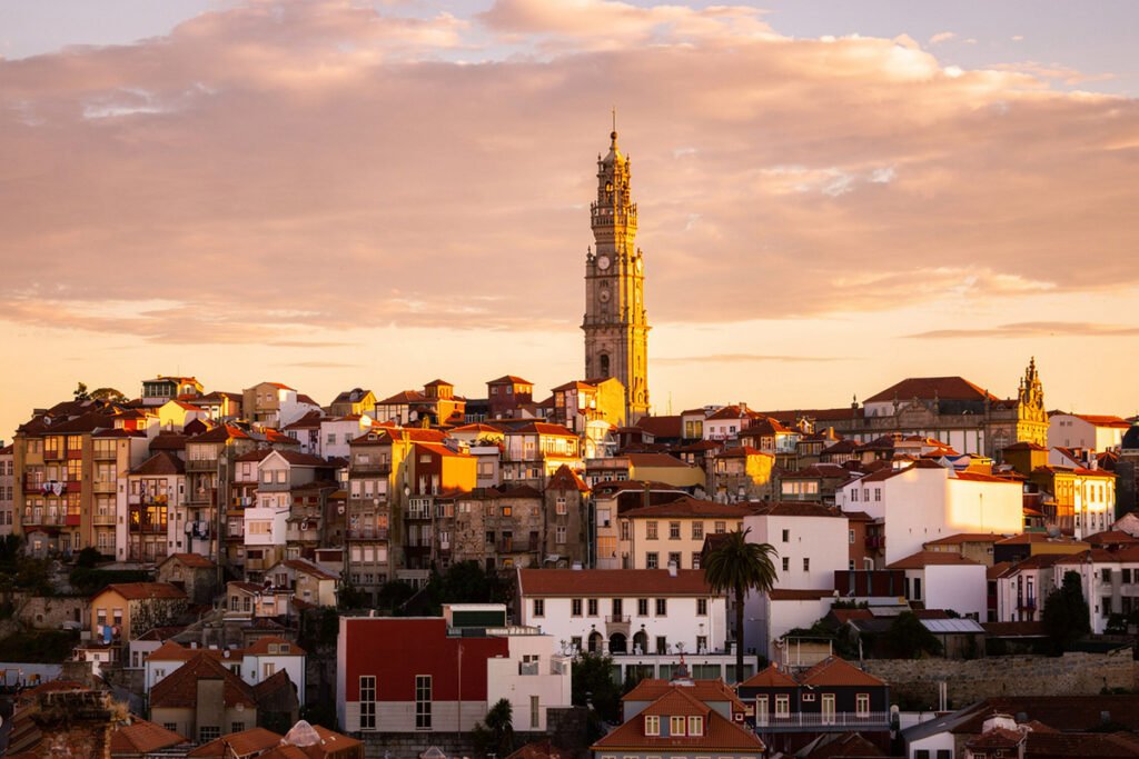 Panoramic view of Porto’s old town at sunset, featuring the Clérigos Tower rising above colorful traditional houses under a golden sky.