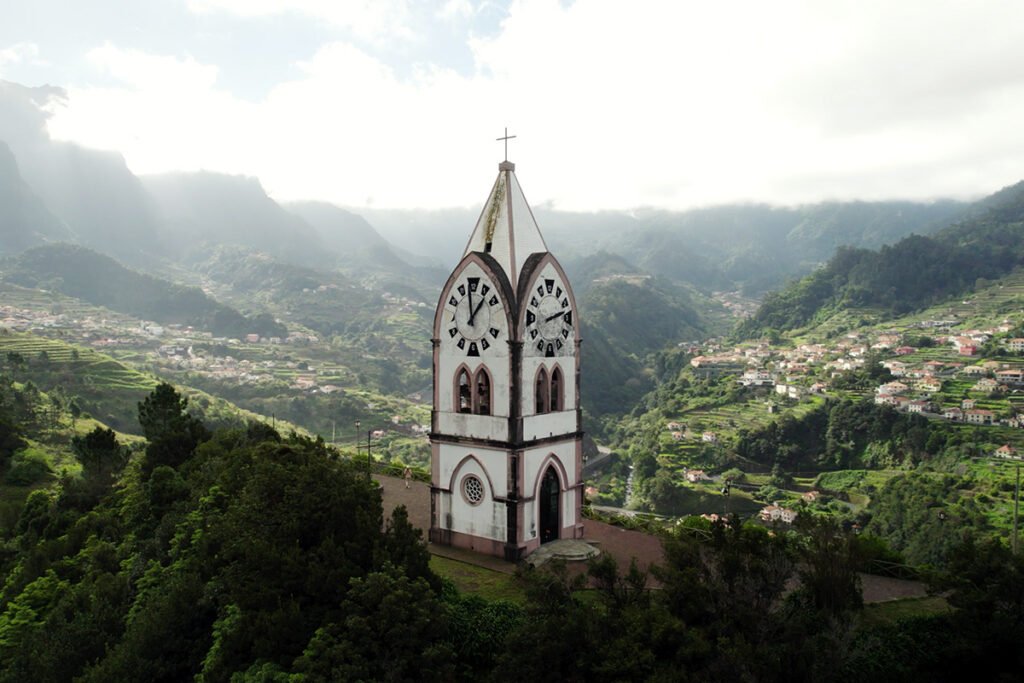 A small white clock tower chapel standing on a green mountain slope overlooking the valley of São Vicente in Madeira