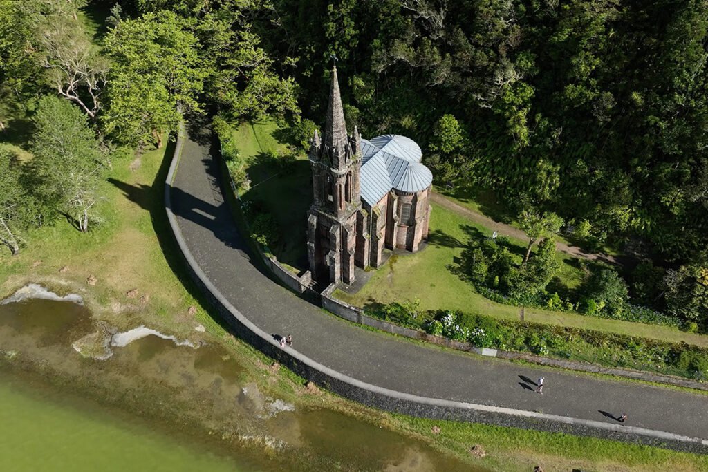 High-angle view of the dark stone Ermida de Nossa Senhora das Vitórias on the shores of Lake Furnas in São Miguel, Azores, surrounded by deep green forest
