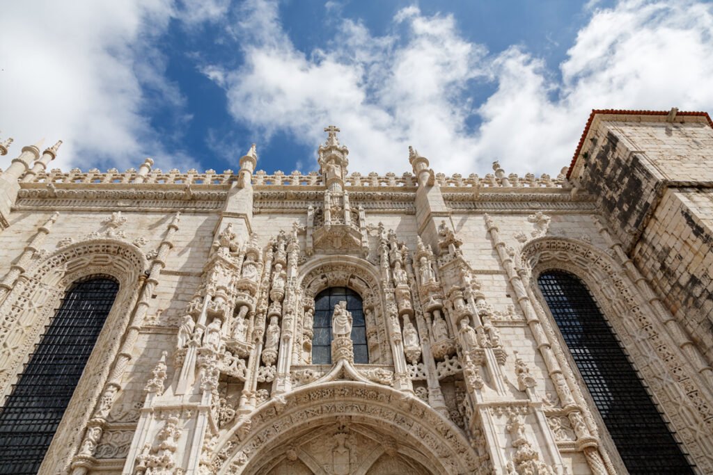 A detailed close-up low-angle view of the highly-ornate limestone facade of the cloister at the Jerónimos Monastery, Lisbon. Intricate Manueline carvings of ropes, sea monsters, and armillary spheres surround tall Gothic arches against a blue cloudy sky