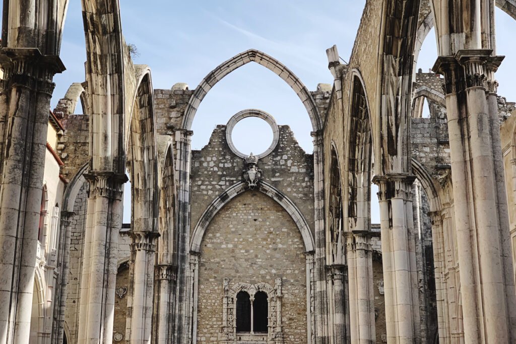 Interior view of the roofless Convento do Carmo in Lisbon, showing the tall, elegant Gothic stone arches reaching toward a clear blue sky