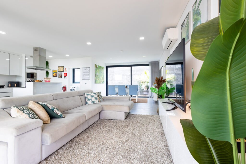 he grand scale open-space living room of a luxury vacation rental in Madeira, featuring an L-shaped sofa, soft carpet, a large TV, and a tropical indoor palm plant