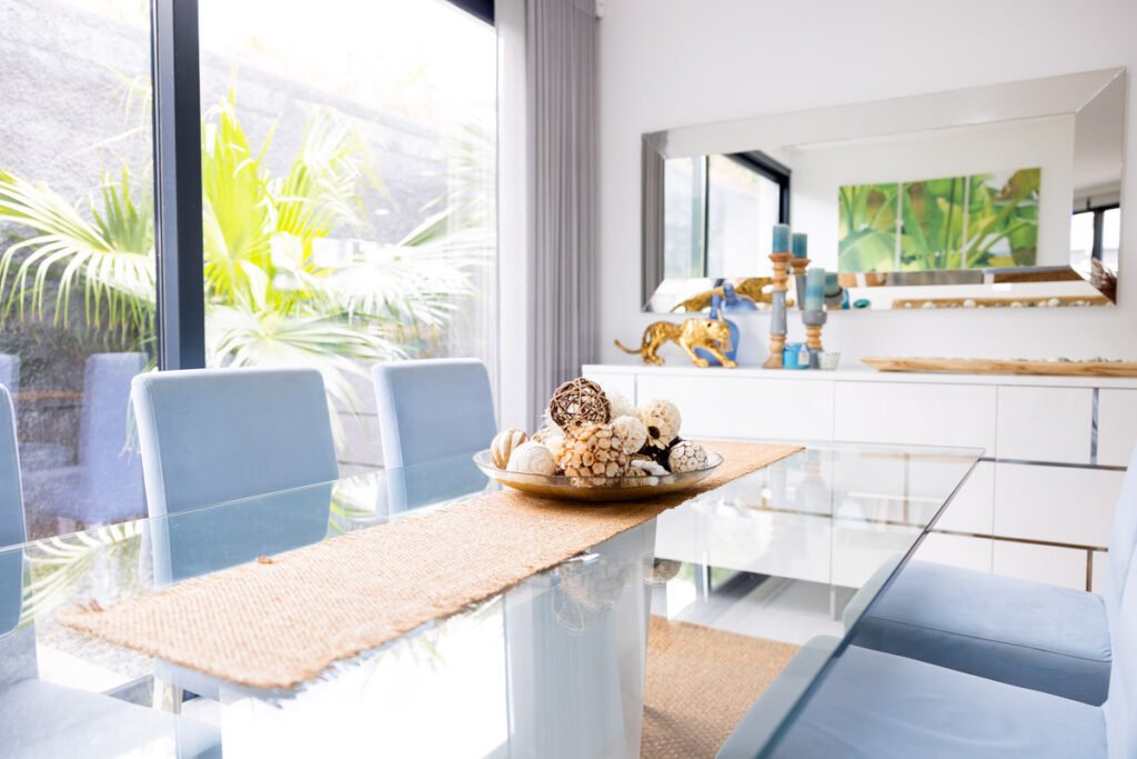 A modern glass-top indoor dining table with elegant gray chairs, reflecting light next to a window in a certified premium AL Caniço property