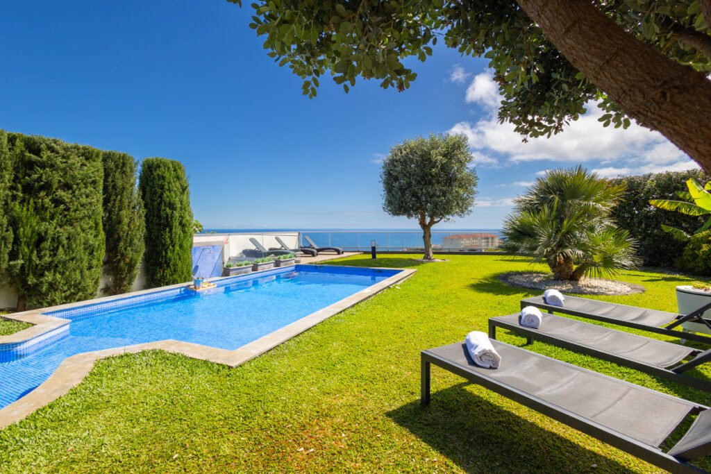 A crisp overhead shot of the private swimming pool and modern sun loungers, surrounded by a lush green lawn in a spectacular house with a view of the sea in Caniço, Madeira