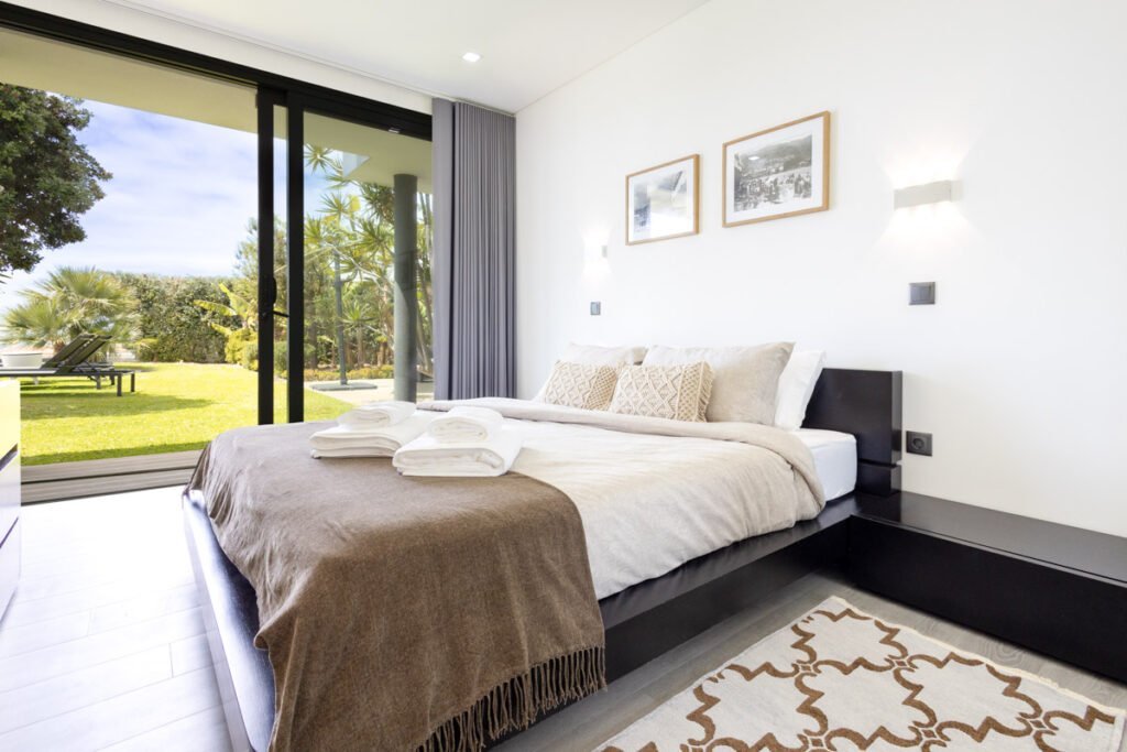 A cozy double bedroom in a luxury house with a view of the sea in Caniço, featuring minimalist decor, a plush brown throw, and a massive glass wall opening to the garden