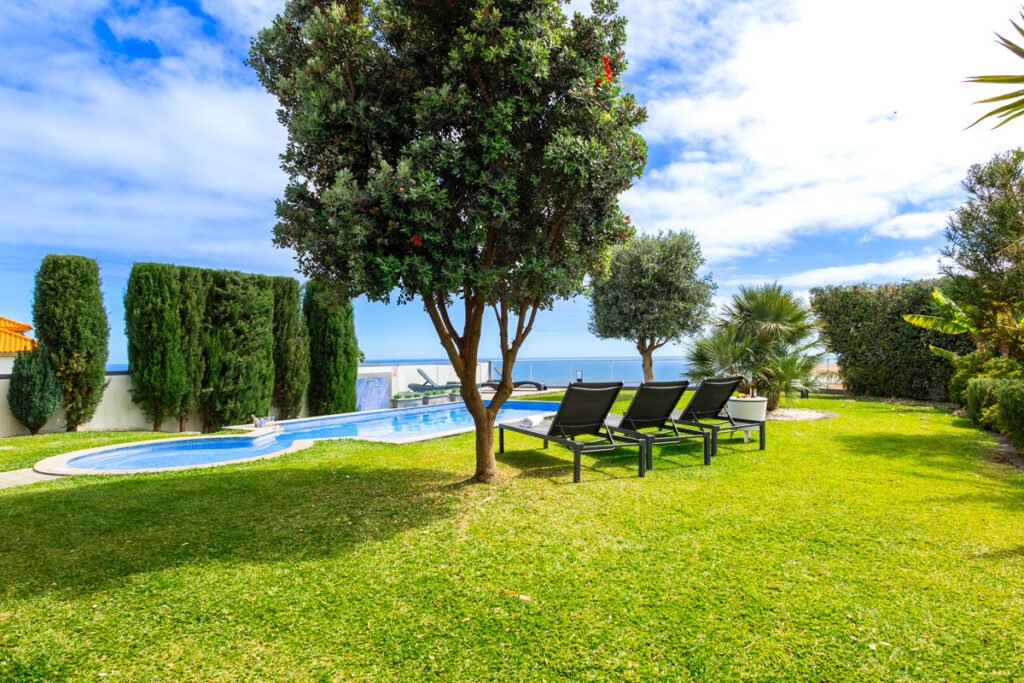 An expansive view of a beautiful beachfront house with a garden facing the sea in Caniço Madeira featuring a large private pool modern sun loungers and tropical trees under a blue sky