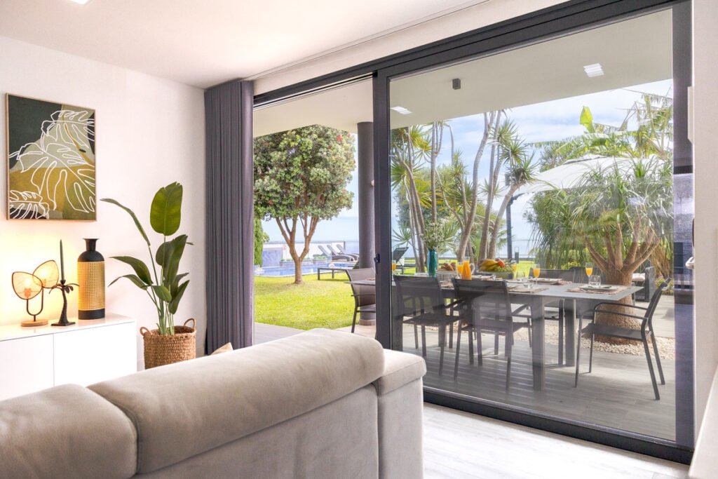 Interior perspective of a luxury vacation rental in Madeira, looking from the cozy living room through open sliding doors toward an al fresco dining table in the seafront garden