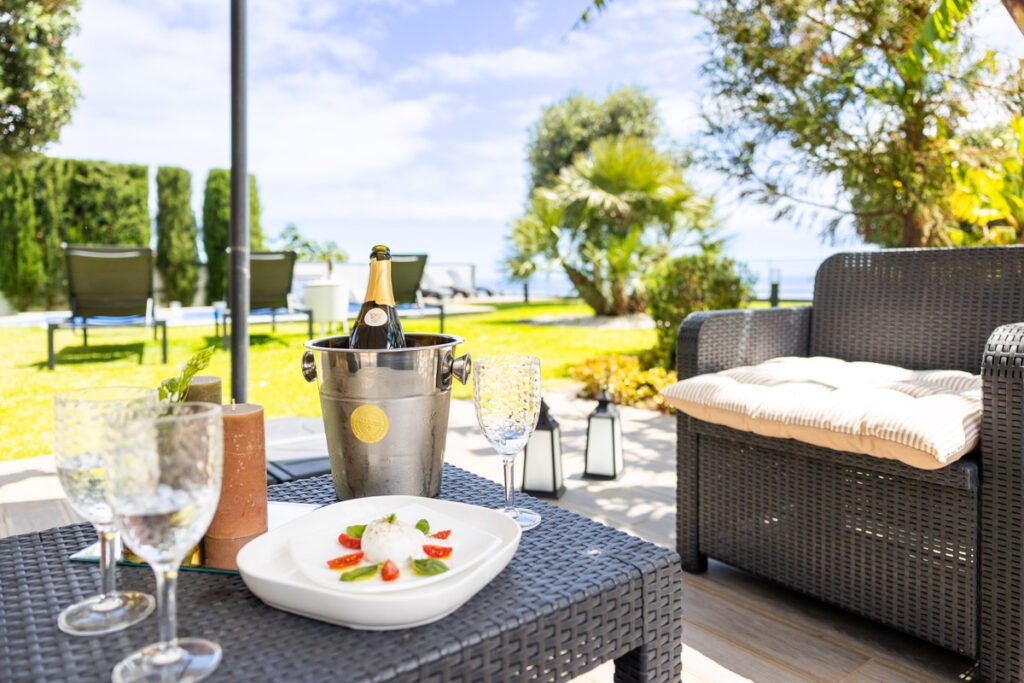 A premium welcome reception on the terrace of a vacation rental in Madeira showcasing a bottle of champagne in an ice bucket and fresh food on a black rattan table with a view of the seafront garden